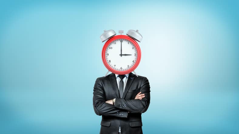 A businessman with crossed arms stands in a front view with a large red alarm clock instead of his head