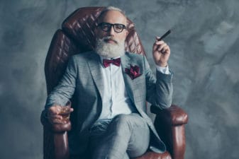 Cool man in glasses, hold cigarette, glass with brandy, in formal wear, tux with red bowtie and pocket square, sit in leather chair over gray background, looking to the camera, shares, stock, money