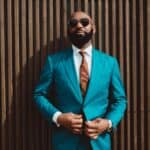 A handsome mature bald bearded black man in a sunglasses and a fashionable blue or teal costume with a tie is standing in front of a wall made of striped wooden timbers and fastening a suit button