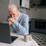 Portrait of pensive bearded senior looking on screen of laptop sitting at table with coffee cup.