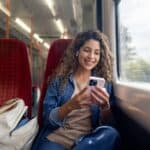 Happy woman commuting on a train and checking her mobile phone while using headphones