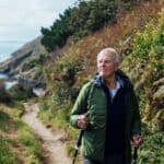 A senior man using hiking poles, on a hike on a coastal path along the coastline of Cornwall. He is looking away from the camera at the view.