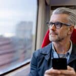 Thoughtful man using his phone while riding on a train and looking through the window