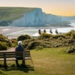 Rear view image depicting a senior man in his 70s sitting on a bench leading down to the iconic Seven Sisters cliffs on the coastline of East Sussex, UK. The man is wearing casual clothing - blue denim jeans, a red checked shirt, navy blue gilet. The man is having a rest from hiking and his hiking pole is leaning up against the bench.