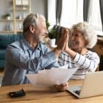 Overjoyed exited middle aged married couple giving high five, finishing doing domestic paperwork together at home. Euphoric happy older mature spouses celebrating successful investment or purchase.
