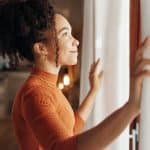 Curtains, happy woman and thinking of future in home, planning and reflection of mindset with view. Window, smile and African girl with vision, ideas and dream for morning inspiration in living room.